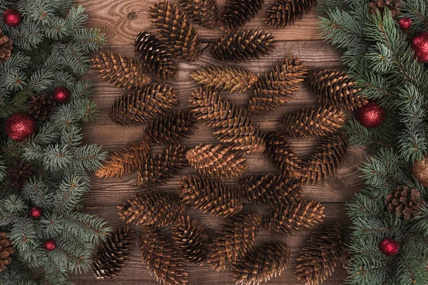 Vista superior de conos de pino marrón con ramitas de abeto y adornos rojos sobre fondo de madera - foto de stock