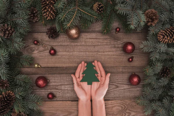 Partial top view of person holding christmas tree symbol above wooden surface with coniferous branches, pine cones and shiny baubles — Stock Photo