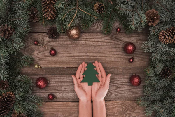 Teilweise Ansicht von oben von Person mit Christbaumsymbol über der Holzoberfläche mit Nadelzweigen, Tannenzapfen und glänzenden Kugeln — Stockfoto