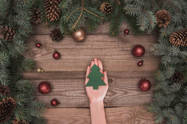 Cropped shot of person holding christmas tree symbol above wooden surface with coniferous branches, pine cones and shiny baubles — Stock Photo