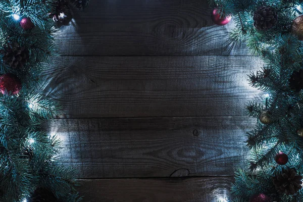 Top view of christmas tree twigs with pine cones, baubles and illuminated garland on wooden background — Stock Photo