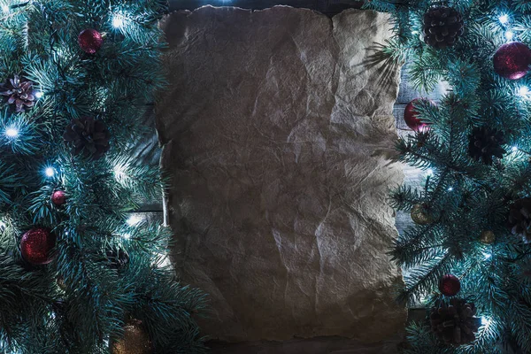 Vista superior de pergamino en blanco y ramas iluminadas de árbol de Navidad con guirnalda, adornos y conos de pino - foto de stock