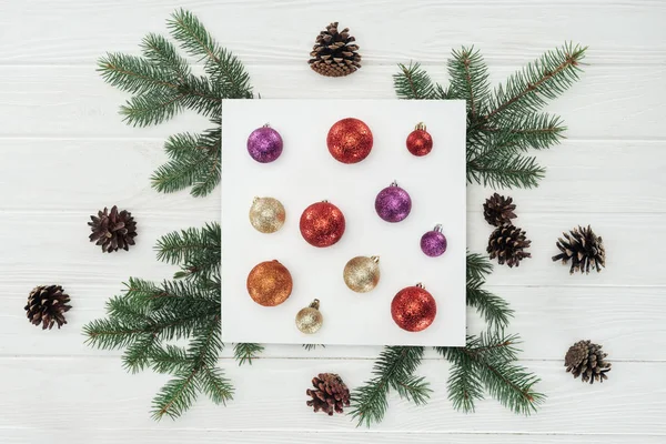 Top view of shiny colorful christmas balls on white card, fir twigs and pine cones on wooden surface — Stock Photo