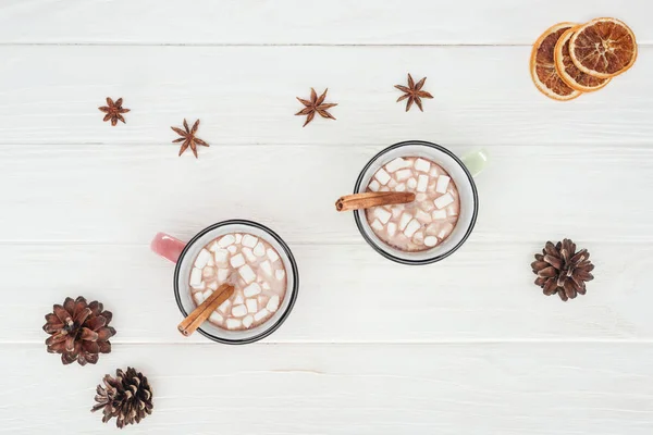 Vista superior de tazas con chocolate caliente y malvaviscos, palitos de canela, anís estrellado y conos de pino en mesa de madera - foto de stock