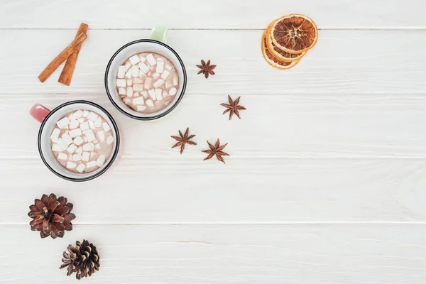 Plat avec des tasses au chocolat chaud et des guimauves, des bâtons de cannelle et des cônes de pin sur une table en bois — Photo de stock