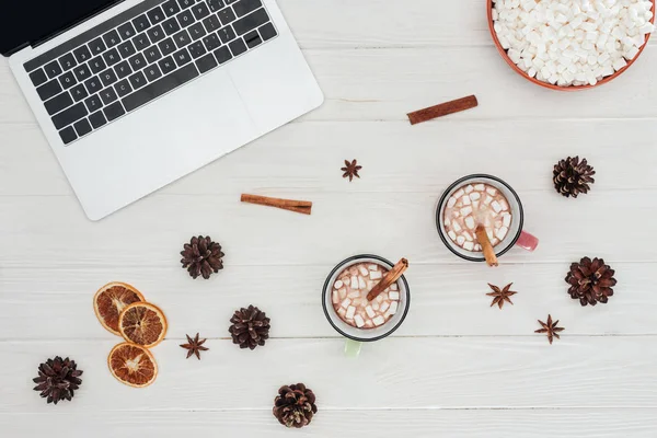 Acostado plano con portátil, tazas de chocolate caliente y palos de canela con malvaviscos en la mesa de madera - foto de stock