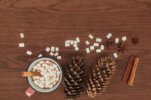 Vista superior da xícara com chocolate quente, marshmallows, cones de pinho e paus de canela na mesa de madeira — Fotografia de Stock