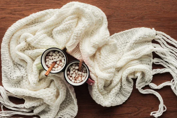 Flache Liege mit Bechern mit heißer Schokolade, Marshmallows und Zimtstangen, weißer Strickschal auf hölzerner Oberfläche — Stockfoto