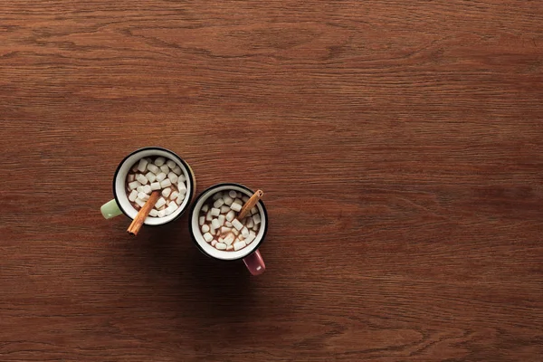 Top view of mugs with delicious hot chocolate, marshmallows and cinnamon sticks on wooden background — Stock Photo