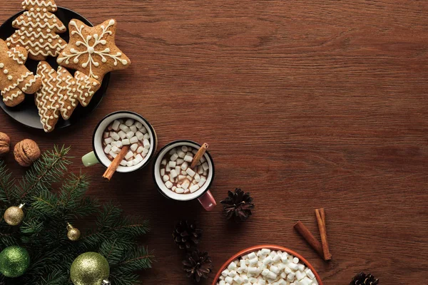 Posa piatta con tazze con cioccolata calda, marshmallow e bastoncini di cannella, gustosi biscotti e ramoscelli di abete con palline lucide sulla superficie di legno, sfondo natalizio — Foto stock