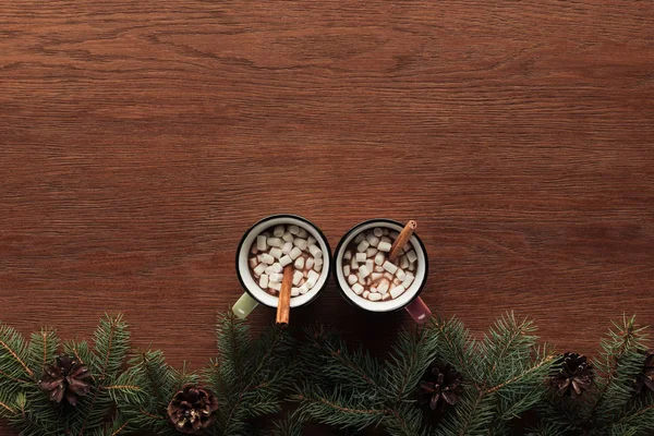 Vista superior de tazas con chocolate caliente, malvaviscos y palitos de canela sobre fondo de madera - foto de stock