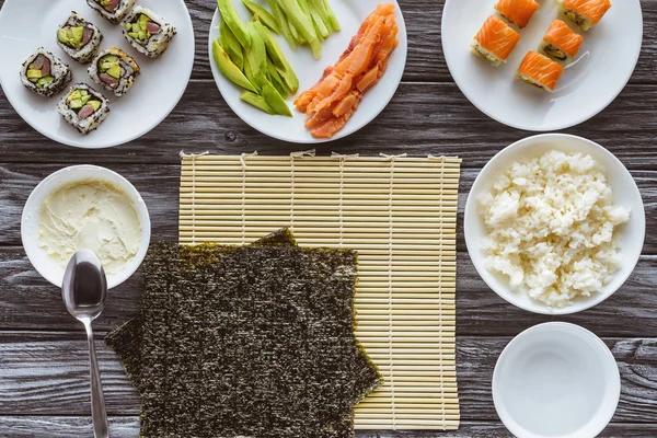 Vista superior de las hojas de nori e ingredientes para preparar delicioso sushi en la mesa de madera - foto de stock