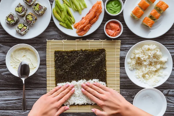 Tiro recortado de la persona preparando sushi con arroz y nori, ingredientes gourmet en la mesa - foto de stock