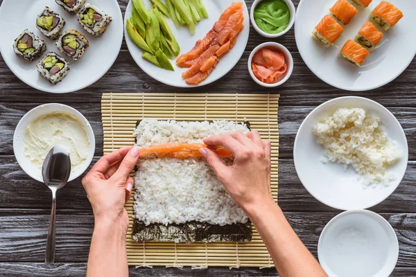 Vue partielle du dessus de la personne qui cuisine de délicieux rouleaux de sushi avec du saumon, du riz et du nori — Photo de stock
