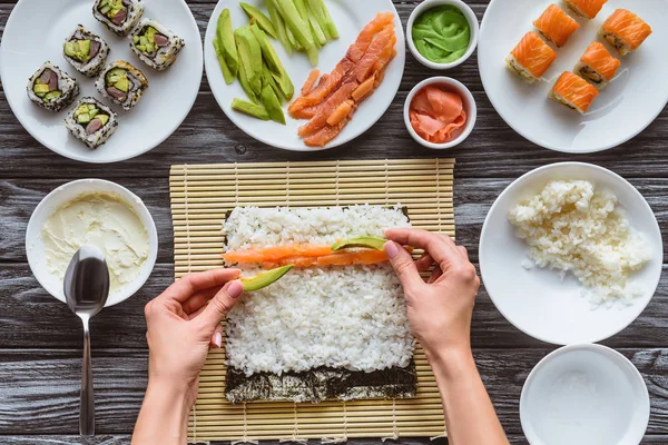 Tiro cortado de pessoa cozinhar delicioso rolo de sushi com salmão, arroz, pepino, abacate e nori — Fotografia de Stock