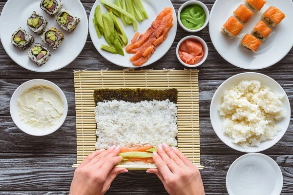 Colpo ritagliato di persona che prepara sushi con riso, nori, salmone e avocado — Foto stock