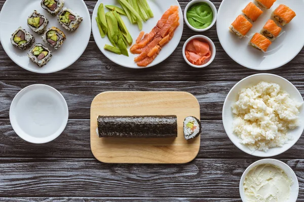 Top view of tasty sushi roll on chopping board and ingredients on wooden table — Stock Photo