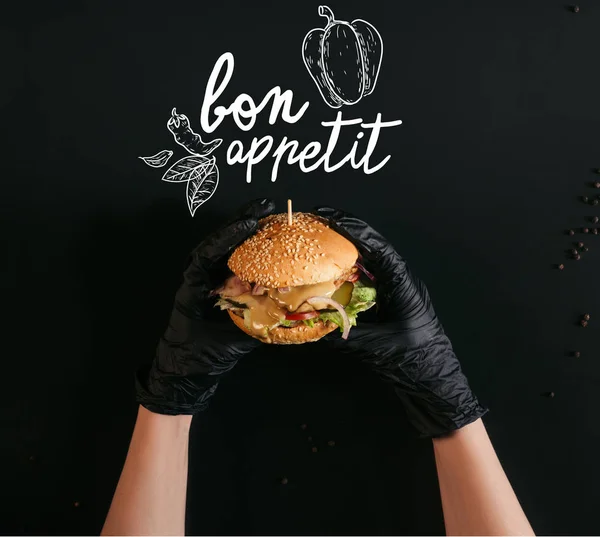 Colpo ritagliato di mani in guanti che tengono delizioso hamburger con tacchino, verdure e Cesare vestirsi di nero con scritte 