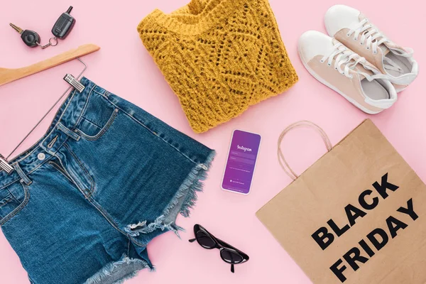 Vista superior de la ropa de moda, teléfono inteligente con aplicación instagram y bolsa de compras con signo de viernes negro - foto de stock