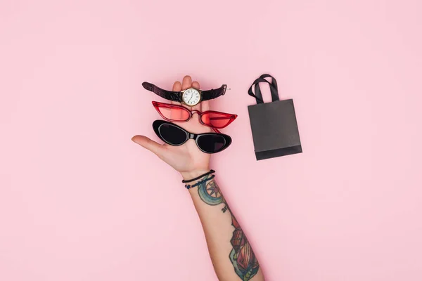 Vista recortada de mujer tatuada sosteniendo gafas de sol de moda y ver cerca de la pequeña bolsa de compras aislado en rosa - foto de stock