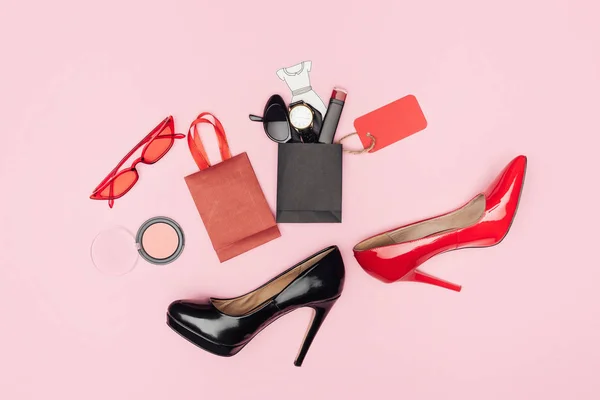 Pose plate avec talons hauts et petits sacs à provisions avec cosmétiques et accessoires isolés sur rose — Photo de stock