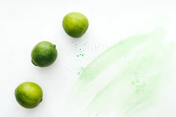 Elevated view of three ripe limes on white surface with green watercolor — Stock Photo