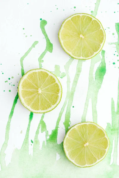 Elevated view of three pieces of ripe limes on white surface with green watercolor — Stock Photo