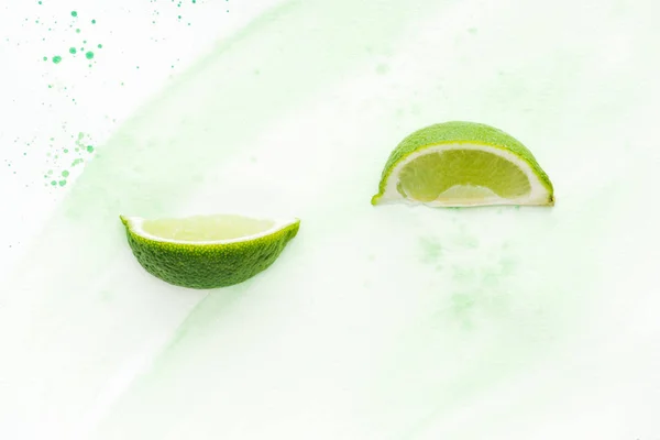 Vue surélevée de deux morceaux de délicieux citrons verts mûrs sur surface blanche avec aquarelle verte — Photo de stock