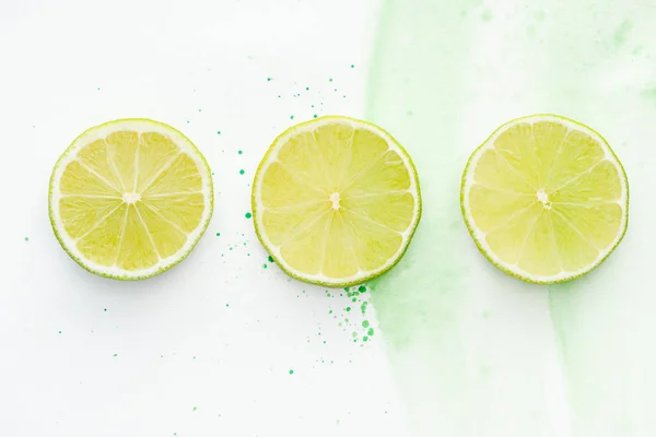 Vue de dessus de trois morceaux ronds de limes mûres sur la surface blanche avec aquarelle verte — Photo de stock
