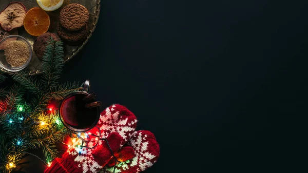 Vista elevada de la copa de vino caliente, guirnalda, manoplas y galletas aisladas en negro, concepto de Navidad - foto de stock