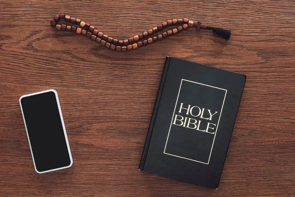 Сверху вид на святую Библию с бусами и смартфоном на деревянном столе — стоковое фото