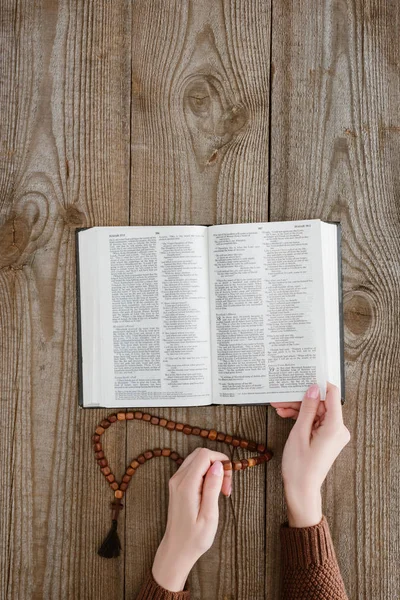Cropped shot of woman reading holy bible and holding beads on wooden table — Stock Photo