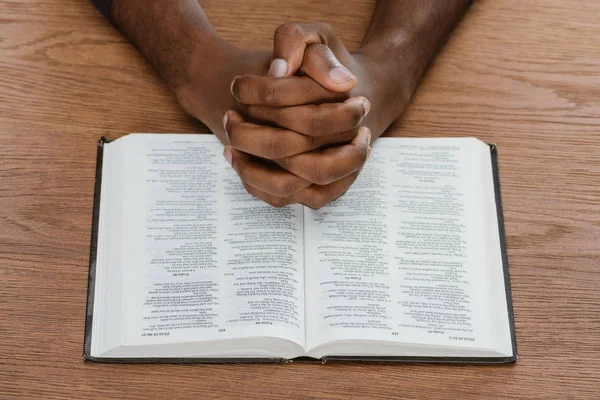 Abgeschnittene Aufnahme eines afrikanisch-amerikanischen Mannes, der mit heiliger Bibel auf hölzerner Oberfläche betet — Stockfoto