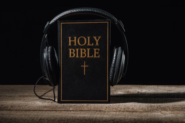 Close-up shot of holy bible with headphones standing on wooden table isolated on black — Stock Photo