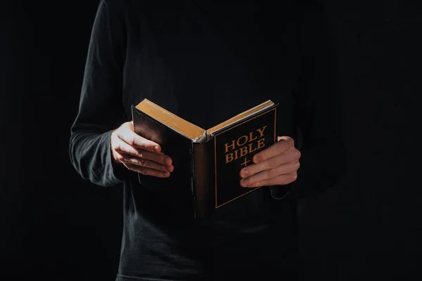 Cropped shot of priest in black cassock holding holy bible isolated on black — Stock Photo