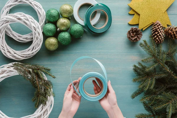 Partial view of woman holding ribbon for handmade christmas wreath decoration on blue wooden tabletop — Stock Photo