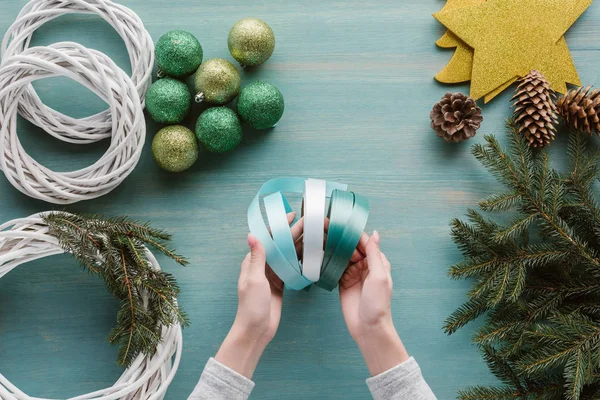 Teilansicht einer Frau mit Schleifen für handgefertigte Weihnachtskranzdekoration auf blauer Holztischplatte — Stockfoto