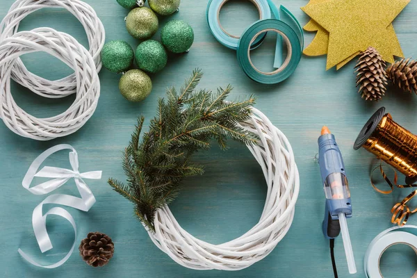 Acostado plano con pistola de pegamento arreglada, juguetes de Navidad y decoraciones para la corona de Navidad hecha a mano en la mesa de madera azul - foto de stock