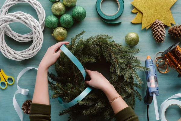 Cropped shot of woman decorating handmade christmas wreath with blue ribbon on blue wooden tabletop — Stock Photo