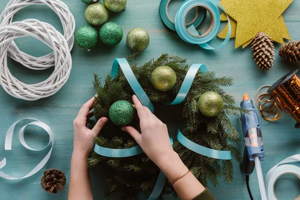 Teilansicht einer Frau, die handgefertigten Tannenbaumkranz mit Weihnachtsspielzeug auf blauer Holzoberfläche dekoriert — Stockfoto