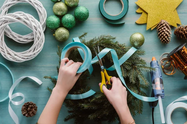 Cropped shot of woman decorating handmade christmas wreath with blue ribbon on blue wooden tabletop — Stock Photo