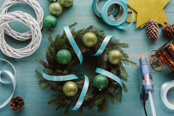 Vista superior de pistola de pegamento arreglada, juguetes chrismtas y decoraciones para la corona de Navidad hecha a mano en la mesa de madera azul - foto de stock