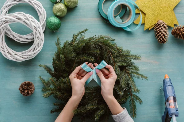 Partial view of woman holding blue ribbon for handmade christmas wreath decoration on blue wooden tabletop — Stock Photo
