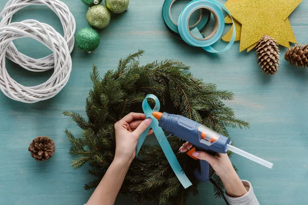 Cropped shot of woman decorating handmade christmas wreath with blue ribbon on blue wooden surface — Stock Photo