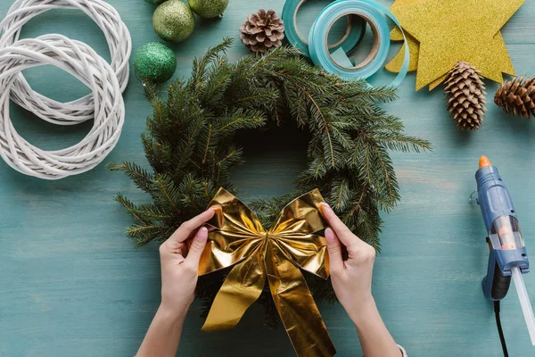 Cropped shot of woman putting golden bow on handmade pine tree wreath on blue wooden surface — Stock Photo