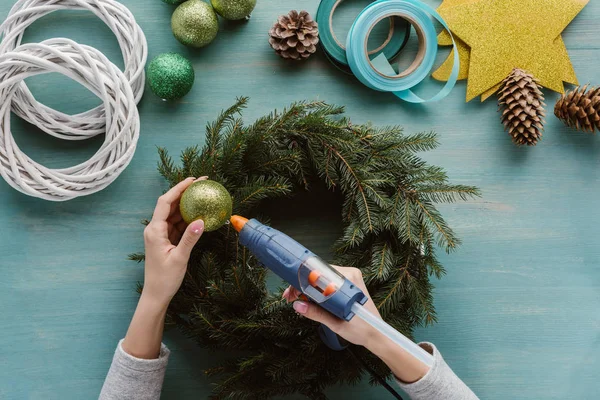 Teilansicht einer Frau, die handgefertigten Tannenbaumkranz mit Weihnachtsspielzeug auf blauer Holzoberfläche dekoriert — Stockfoto