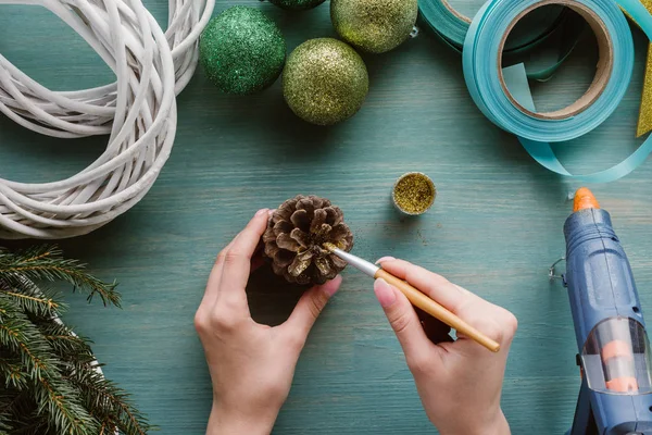 Partial view of woman coloring pine cone with golden paint on blue wooden surface — Stock Photo