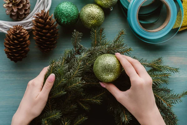 Partial view of woman decorating handmade pine tree wreath with christmas toys on blue wooden surface — Stock Photo