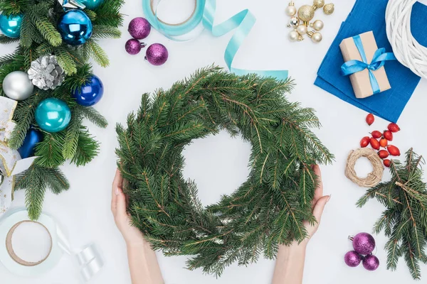 Partial view of woman holding handmade pine tree wreath at tabletop with chrismtas decorations — Stock Photo