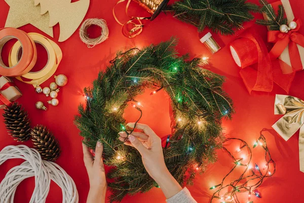 Partial view of woman decorating handmade christmas wreath with lights on red background — Stock Photo